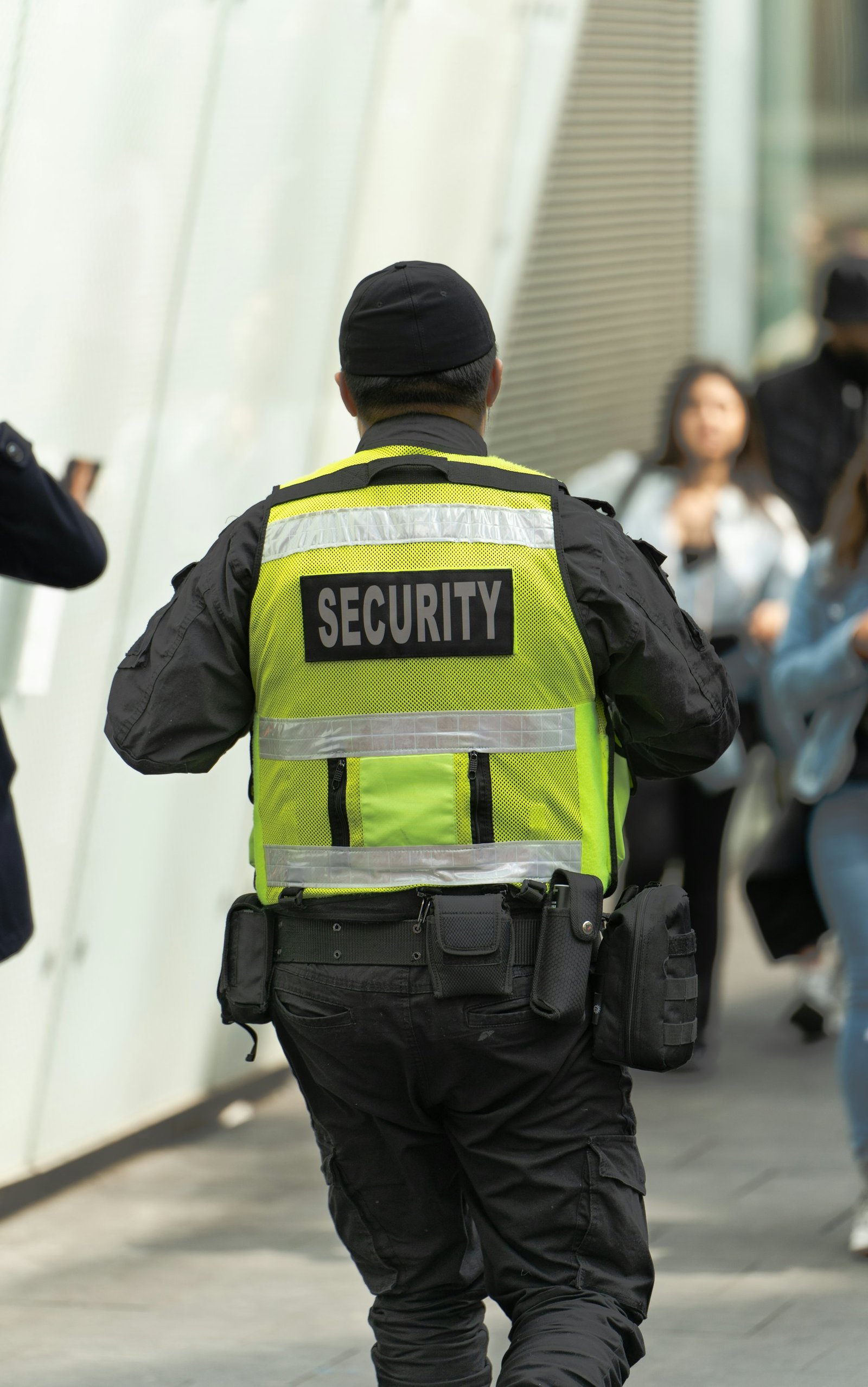 Security guard in a hi-vis vest marked SECURITY, seen from behind walking along a building frontage with pedestrians nearby